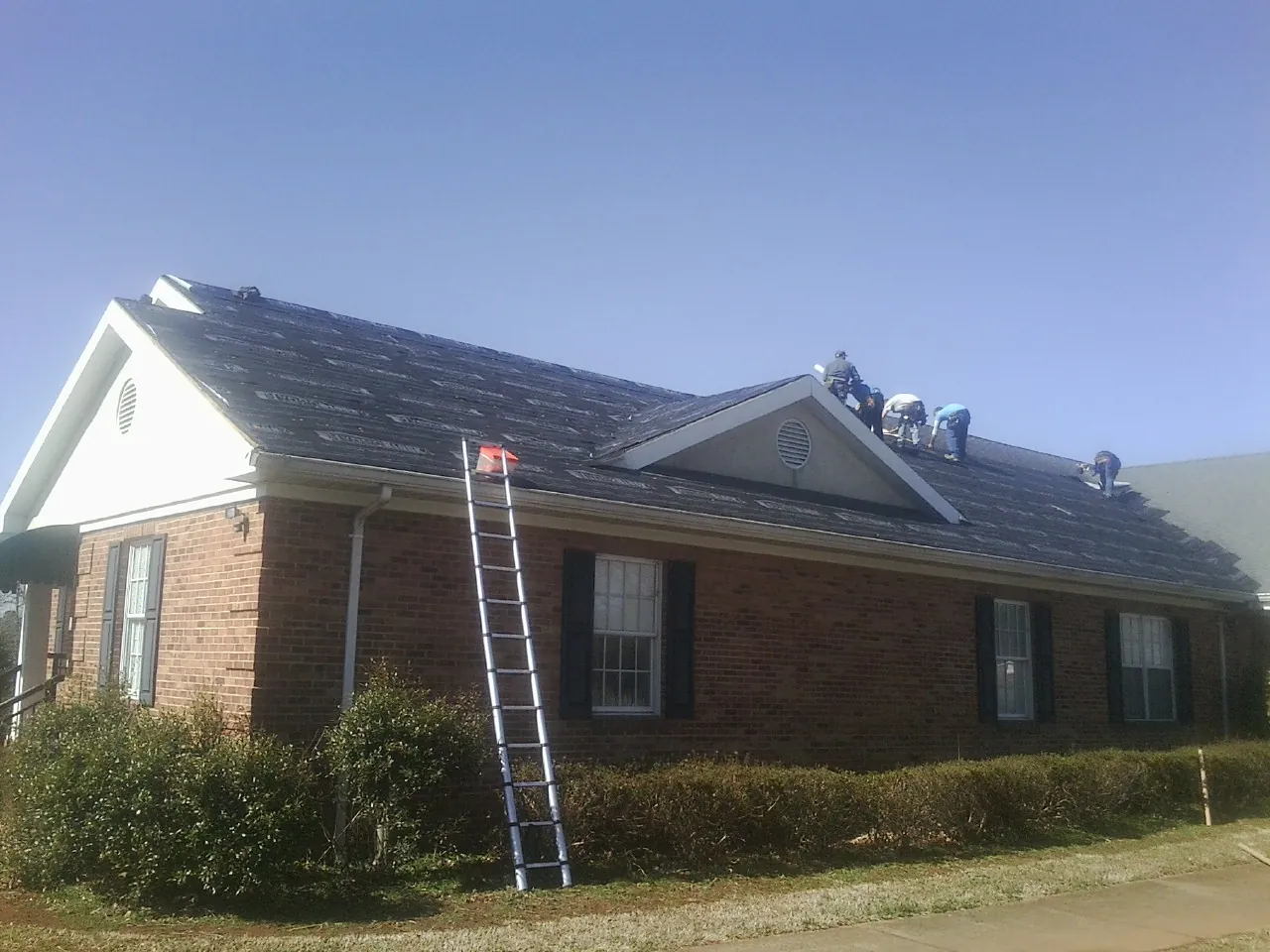 Crew tearing shingles off a brick ranch roof with a ladder set — Emergency Roof Repair work by our crew in Advent