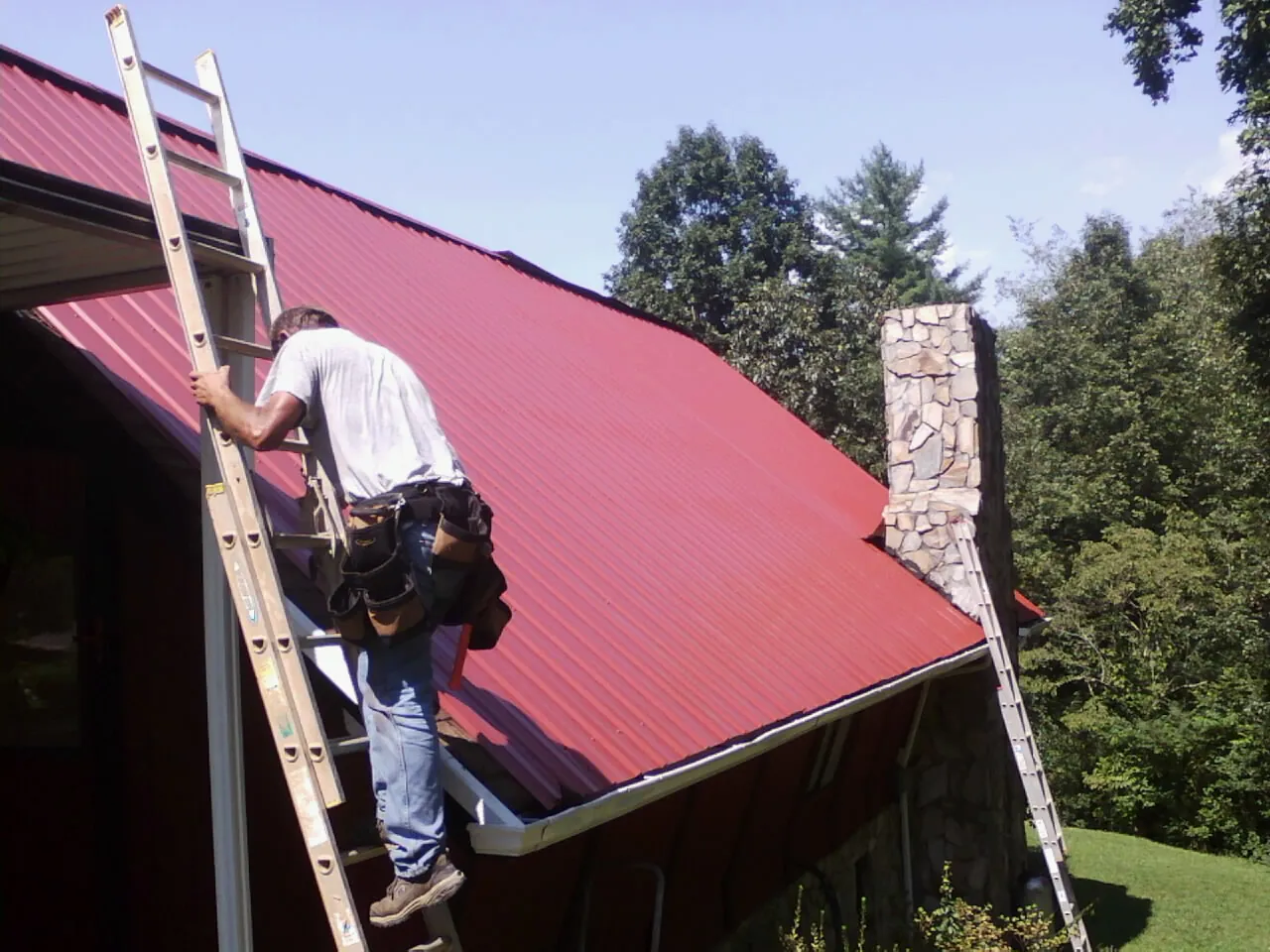 Roofer on a ladder beside a red metal roof and stone chimney — Soffit & Fascia Repair by Roofing Brothers in Advent